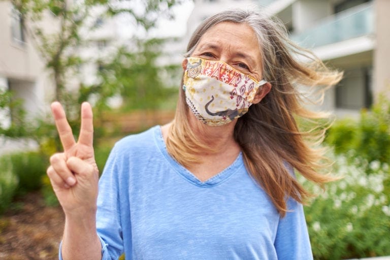 An older woman smiles at the camera with a face mask or protective mask and forms the peace sign with her fingers as an alternative greeting