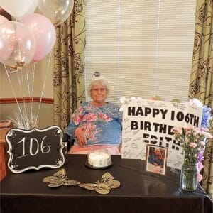 ASC Centenarian Edith Hallett wearing tiara with "106" on it and sitting in front of a table with pink and white balloon and sign that reads "Happy 106th Birthday Edith"