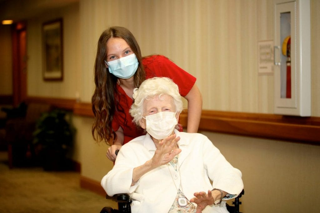 CNA wearing a mask pushing the wheelchair of a senior woman who is also wearing a mask.