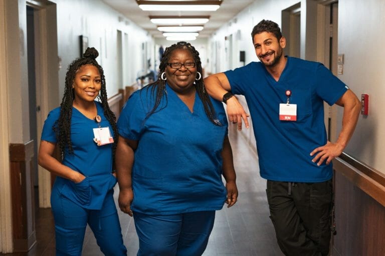 Three nurses in blue scrubs casually standing in a medical setting hallway.