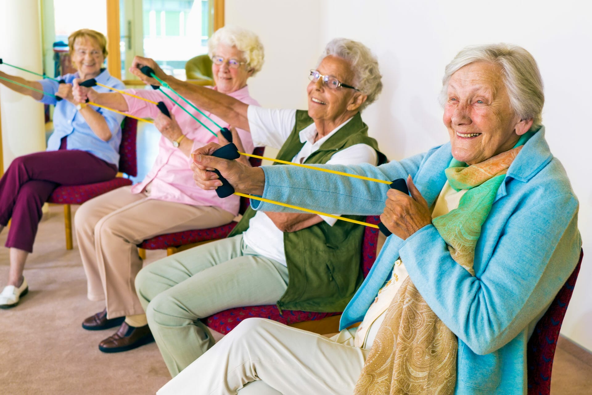 Group of four smiling senior women toning their arms with elastic strengthening bands while seated in fitness class.