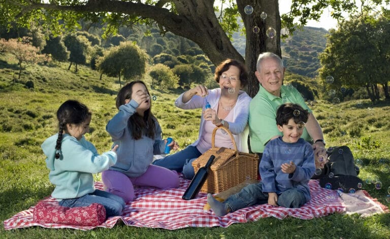 Two grandparents and three grandchildren enjoying a picnic on a grassy hillside.