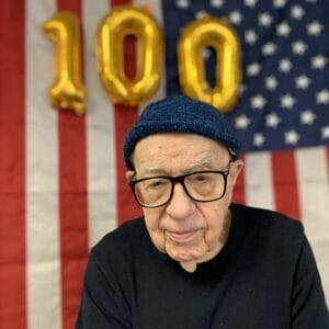 Centenarian Chester Kozlowski behind American flag backdrop with three golden balloons that spell out the number "100"
