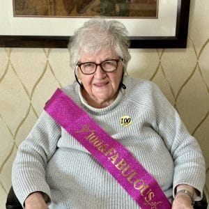 Centenarian Maxine McNabb in a gray sweater with a pink sparkly sash that reads "100 and Fabulous" in metallic gold lettering