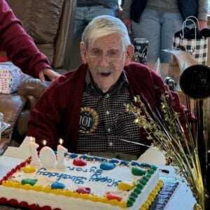 Centenarian Byron "Pop" Wadsworth in red cardigan and black and white plaid button down shirt with a ribbon pinned to his shirt that reads "101st Birthday" sitting in front of a birthday cake that has the number candles "101" on it