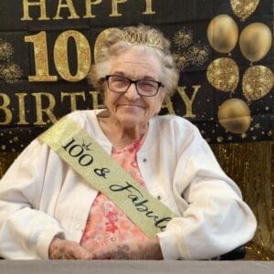Centenarian Betty Jean Hall dressed in pink blouse and white cardigan with a gold sash that reads "100 & Fabulous" with a black and gold "Happy 100th Birthday" banner in the background