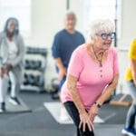 A small group of diverse seniors work out in a small fitness studio together. They are each dressed comfortably as they hold a stretch and focus on their breathing.
