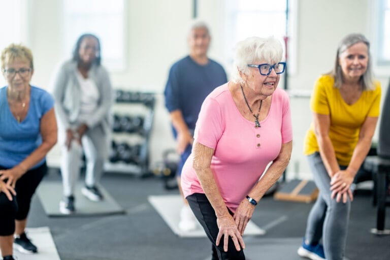 A small group of diverse seniors work out in a small fitness studio together. They are each dressed comfortably as they hold a stretch and focus on their breathing.