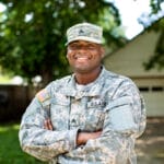 African American Sergeant U.S. Army standing arms crossed in front of residential home and smiling.