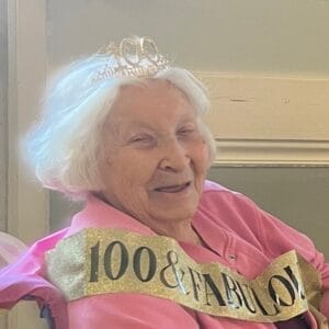 Centenarian Delores "Peggy" Burns in a pink sweater with a gold sash that reads "100 & Fabulous" and wearing a crown