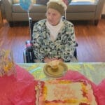 Centenarian Virginia "Ginny" Knight wearing a blue and white floral blouse and white turtle neck while sitting in front of her pink, magenta, yellow and white floral birthday cake that reads "Happy Birthday Virginia"