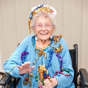 Centenarian Doris Gagnon sitting in a wheelchair with a blue sweater and "Birthday Girl" crown with colorful celebratory confetti draped on her head and sweater
